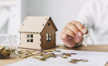 Persona entregando llaves junto a una casa en miniatura y monedas sobre la mesa, simbolizando una inversión inmobiliaria.