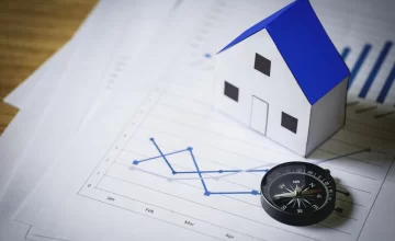 Modelo de casa con techo azul y brújula sobre gráficos financieros, representando análisis de plusvalía inmobiliaria y valorización de propiedades.